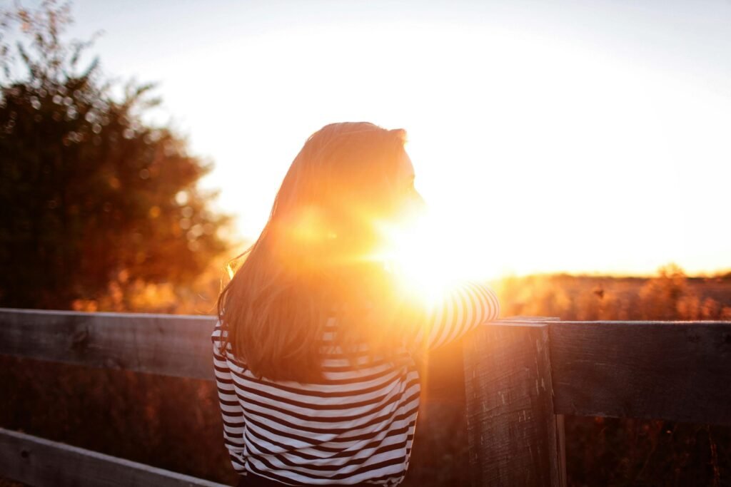 A woman leans against a wooden fence, basking in the warm glow of a sunset on a rural farm.