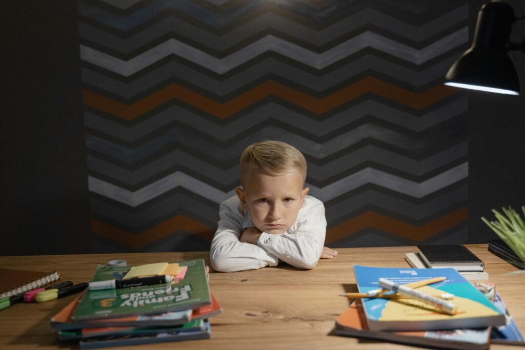 pexels-photo-5303548-5303548 A young boy leaning on a table surrounded by books and coloring materials, looking unhappy with studying.